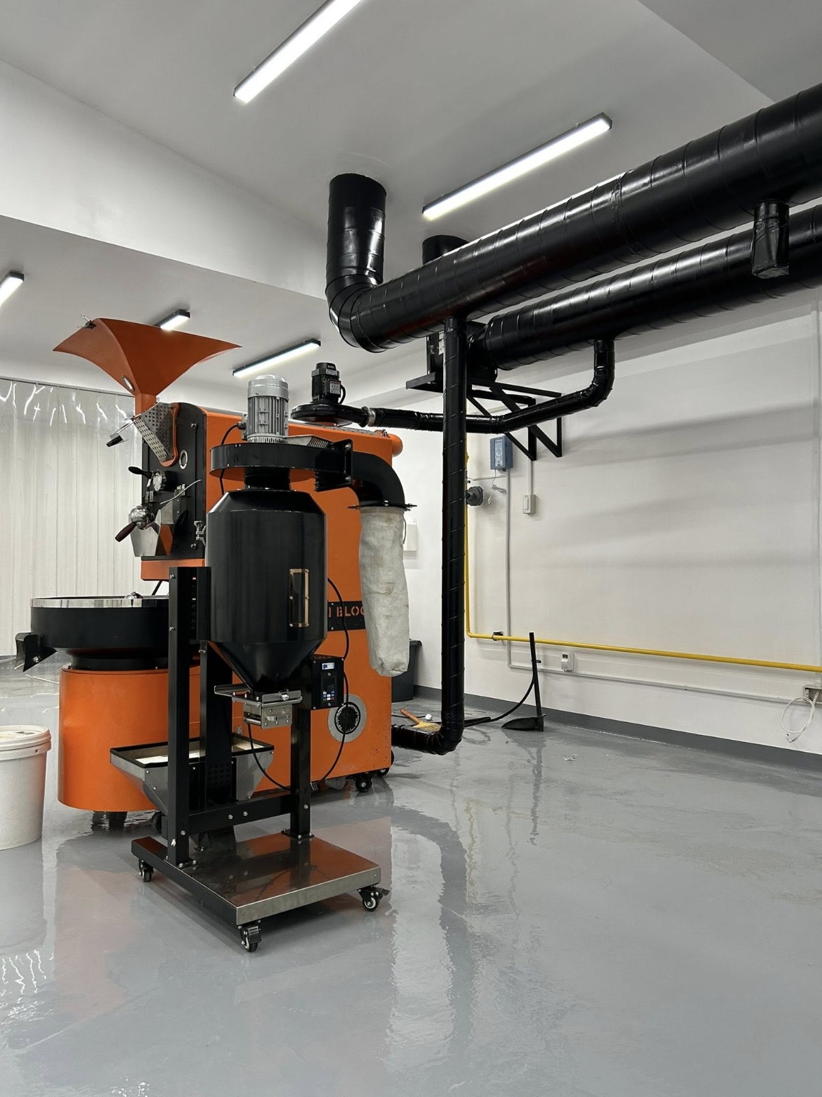 Commercial coffee roastery - orange drum roaster with cyclone and dust collector unit. Yellow natural gas supply line visible along the right wall in the background.